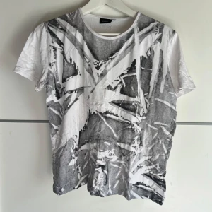 Vit och grå t-shirt med grafiskt tryck Lindex - Cool t-shirt från Lindex i vitt med grått grafiskt tryck på framsidan. Klassisk rund hals och korta ärmar. Skön och lätt i bomull, perfekt till jeans eller shorts för en avslappnad vibe.