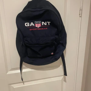 Mörkblå ryggsäck från GANT - Säljer en stilren mörkblå ryggsäck från GANT Sportswear. Väskan har ett stort huvudfack, en praktisk ytterficka med dragkedja och justerbara axelband. Materialet är slitstark polyester och loggan syns tydligt framtill. Perfekt för skola eller cityhäng. Har använt den några gånger men är som ny