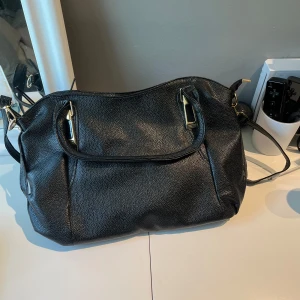 Svart axelväska i skinn med dragkedja - Snygg svart axelväska i skinn med guldiga metalldetaljer. Väskan har flera fack inuti, inklusive ett med dragkedja, och gott om plats för allt du behöver. Perfekt för dig som vill ha en stilren och rymlig väska till vardagen.