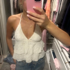 Vit volangig linnetopp med knyt - Supersöt vit linnetopp med volanger och knyt i nacken. Toppen är ärmlös och har en djup v-ringning framtill samt öppen rygg. Perfekt för varma dagar och ger en luftig och fräsch känsla. Materialet är lätt och har en skrynklig struktur som ger extra liv till looken.