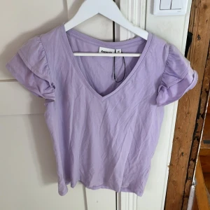 Ljuslila topp med volangärm Kappahl XS - Supersöt ljuslila topp från Kappahl i storlek XS. Toppen har v-ringning och korta volangärmar som ger en extra touch. Tillverkad i mjuk bomull, perfekt för vår och sommar. Enkel att matcha med jeans eller kjol för en chill look.