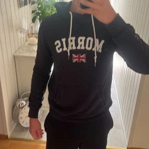 Svart hoodie från Morris med flagga - Mörkblå hoodie från Morris med vit broderad logga och brittisk flagga på bröstet. Tröjan har huva med vita snören och en stor magficka. Tillverkad i mjuk bomull och har långa ärmar. Perfekt för en chill och stilren look. Hör gärna av om ni har frågor 