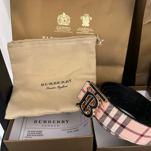 Rutig Burberry bälte - Snyggt Burberry bälte i klassiskt rutigt mönster med rosa, beige och bruna toner. Bältet har ett stort B-spänne i svart och guld som ger en lyxig känsla. Tillverkat i skinn och levereras med originalpåse och box. Perfekt accessoar för att lyfta din outfit.