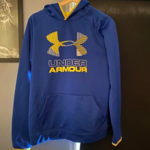 Blå Under Armour träningshoodie  - Snygg blå hoodie från Under Armour med stor gul och vit logga på bröstet. Tröjan har huva med gul insida, känguruficka och gula detaljer vid ärmsluten. Perfekt för dig som gillar sportig stil och vill sticka ut lite extra.