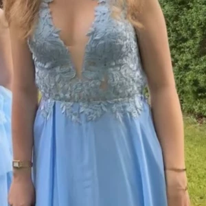 Ljusblå lång-bal-klänning med spetsdetaljer - Superfin ljusblå långklänning med djup v-ringning och blommiga spetsdetaljer upptill. Klänningen har smala axelband, omlottkänsla och flowy kjol i tunt tyg. Perfekt för bal eller student! Säljes då den inte längre kommer till användning.