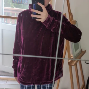 Mörklila sammetströja med snörning - Mysig mörklila tröja i sammet med hög krage och snörning nertill. Långärmad och loose fit, perfekt för dig som gillar en chill och trendig stil. Materialet är mjukt och glansigt vilket ger en lyxig känsla. Den är stor och rymlig och perfekt för en mysig kväll hemma.