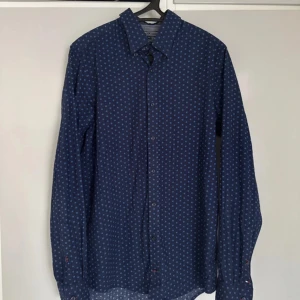 Mönstrad blå skjorta Tommy Hilfiger M - Snygg mörkblå skjorta från Tommy Hilfiger i slim fit med små blommiga mönster i ljusblått. Skjortan har långa ärmar, klassisk krage och knäppning framtill. Tillverkad i mjuk bomull och har diskreta logodetaljer vid manschetten.