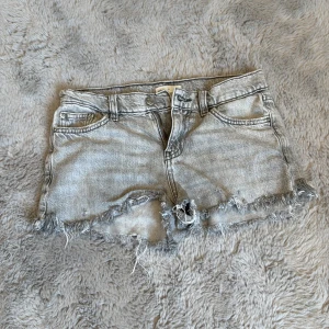 Grå jeansshorts med fransig kant - Snygga grå jeansshorts med rå, fransig kant och klassisk femficksdesign. Shortsen har låg midja och slitna detaljer för en avslappnad vibe. Perfekta för sommardagar och festivaler. Passar dig som gillar en cool och avslappnad stil.