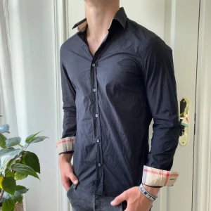 Svart Burberry skjorta - Riktigt tuff skjorta ngl. Gillar hur mönstret är på ärmarna och kragen! Modellen är 180,75 kg och bär S. Känns som en slim fit. Inga defekter! 
