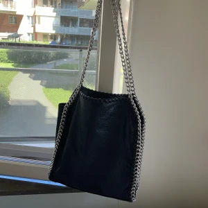 Svart tiamo axelväska med kedjedetaljer  - Snygg svart axelväska från Tiamo väldigt ny finns bara en liten fläck på insidan men jag tror att man kan ta bort med tvål och vatten .