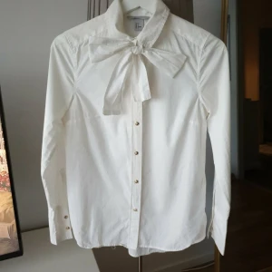 Vit skjorta med rosett och guldknappar - Stilren vit skjorta från H&M med lång ärm, snygga guldknappar och en stor rosett vid kragen som ger en extra touch. Skjortan är rak i modellen och tillverkad i bomull, perfekt för dig som gillar klassiska plagg med en twist. Notera 2 små märken på atm och rosetten, se bild