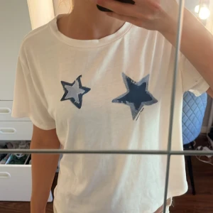 Vit t-shirt med blå stjärnor - Vit t-shirt i bomull med två tryckta stjärnor i blått och mörkblått framtill. Klassisk rund halsringning och korta ärmar. 