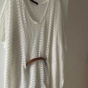 Vit stickad blus med bälte - Vit, luftig stickad blus från Selected Femme med korta ärmar och v-ringning. Kommer med ett flätat brunt bälte i midjan som ger en snygg siluett. Perfekt för varma dagar och enkel att styla över linne eller bikini.