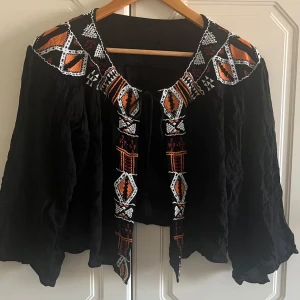 Svart bolero  med broderier och snörning - Unik svart bolero med broderade detaljer i orange, vitt och brunt runt kragen och framtill. Boleron har vida ärmar och knytband med tofsar framtill. Perfekt för dig som vill sticka ut med coola mönster och boho-vibe.