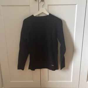 Svart ribbad tröja Peak Performance - Svart långärmad tröja från Peak Performance i ribbad stickad design. Klassisk rund halsringning och diskret logga nertill. Perfekt för dig som gillar en enkel men stilren look. Materialet känns mjukt och passar bra till lager-på-lager.