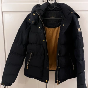 Mörkblå pufferjacka från Morris Lady - Mörkblå pufferjacka från Morris Lady med guldiga detaljer och dragkedjor. Jackan har huva, två fickor med dragkedja och snyggt emblem på ärmen. Även en ficka på insidan av jackan med dragkedja. Insidan är beige och jackan är perfekt för kalla dagar. Klassisk och stilren modell som håller dig varm. Denna jacka kommer från Morris Lady, en numera nedlagd damkollektion, vilket gör den till en sällsynt modell. Storlek: small. 