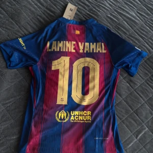 FC Barcelona Lamine Yamal matchtröja - Barça matchtröja med Lamine Yamal 10 på ryggen. Klassiska blå och röda ränder, officiella klubbmärket och Nike-logga framtill. Sponsorer som Spotify och UNHCR tryckta på tröjan. Tillverkad i lätt, ventilerande funktionsmaterial.