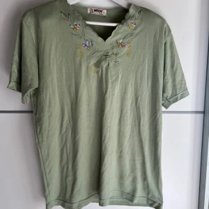 Grön blus med broderi från MEEN - Söt ljusgrön blus från MEEN med korta ärmar och v-ringad hals. Halsringningen har broderade blommor i flera färger och fina blad, vilket ger en unik och vintage-inspirerad look. Tillverkad i mjuk bomull, perfekt för dig som gillar detaljer.