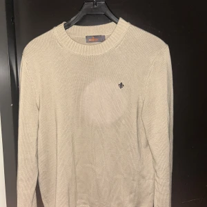 Beige stickad tröja Morris - Beige stickad tröja från Morris med rund halsringning och diskret broderad logga på bröstet. Tröjan har lång ärm och ribbade muddar vid hals, ärmslut och nederkant. Perfekt för lager-på-lager och enkel att matcha med jeans eller chinos.