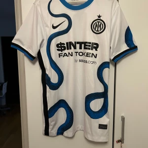 Inter Milan vit bortatröja Nike - Inter Milans officiella bortatröja i vitt med blå och svarta detaljer. Tröjan har ett unikt ormmönster i blått och svart, klubbmärke på bröstet och sponsortryck. Tillverkad i lätt och ventilerande material, perfekt för match eller träning.