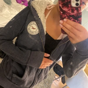 Blå teddyjacka från Hollister XS - Mysig blå teddyjacka från Hollister med vit fluffig insida och huva. Jackan har dragkedja framtill, två fickor och en rund Hollister Los Angeles-logga på bröstet. Perfekt för kyliga dagar när du vill vara både bekväm och snygg.
