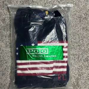 Mörkblå stickad tröja Polo Ralph Lauren - Mörkblå stickad tröja från Polo Ralph Lauren med amerikansk flagga framtill och RL-broderi. Tröjan har rund hals, ribbade muddar och lång ärm. Perfekt för dig som gillar klassisk och preppy stil.