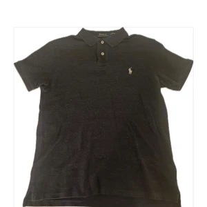Svart pikétröja Polo Ralph Lauren M - Klassisk svart pikétröja från Polo Ralph Lauren i slim fit. Tröjan har korta ärmar, krage med knappar och den ikoniska broderade loggan på bröstet. Tillverkad i 100% bomull för en skön och luftig känsla.