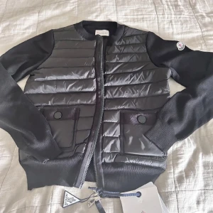 Cardigan från Moncler - Snygg svart pufferjacka från Moncler med quiltad front och stickade ärmar. Jackan har rund hals, två fickor med knappstängning och dragkedja framtill. Perfekt för dig som vill ha en stilren och sportig look med exklusiv känsla.