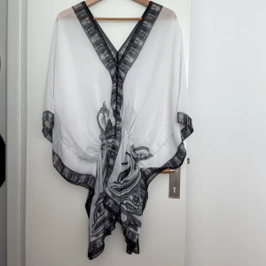 Vit och svart kaftan med mönster - Luftig, see-trough kaftan i vitt med svart och grått paisleymönster. V-ringning fram och bak, fram med dekorativa knappar och breda, lösa ärmar. One size. Resår i midjan. 
