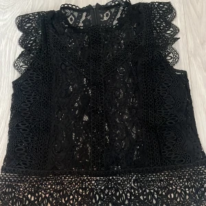 Svart spetsblus från Zara - Supersnygg svart blus från Zara i storlek XS, helt i spets med vackra broderade detaljer och volang längs axlarna. Blusen är ärmlös och har hög hals, perfekt för att lyfta vilken outfit som helst. Dragkedja i ryggen för enkel påklädning.