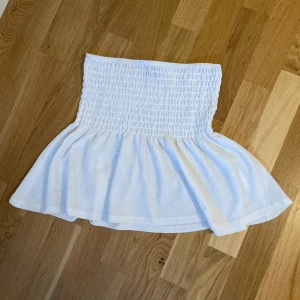 Vit ärmlös smockad topp från Gina tricot - Supersöt vit topp med smockad överdel och volangig nederdel. Ärmlös och axelbandslös modell som sitter tajt upptill och faller ut i en luftig volang. Perfekt för varma dagar och enkel att matcha med shorts eller kjol.