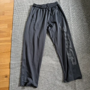 Svarta mjukisbyxor från Gasp - Svarta mjukisbyxor från Gasp med stort tryck längs ena benet. Byxorna har resår och snörning i midjan samt två sidofickor. Perfekta för gymmet eller chill hemma, med en loose fit och sportig vibe.