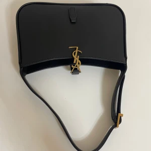 Svart axelväska från Saint Laurent - Snygg svart axelväska från Saint Laurent i slätt skinn med guldigt YSL-spänne framtill. Väskan har en stilren, rektangulär form och justerbar axelrem med gulddetalj. Perfekt för dig som gillar klassisk och lyxig design med en modern touch.