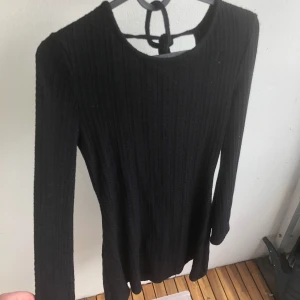 Svart ribbad stickad kortklänning - Superfin ribbadt mönster kort klänning i svart med lång ärm och snygg öppen rygg med knytband. Materialet är mjukt och stretchigt, stickad med tunnare tyg perfekt som klänning på hösten.  (Har lagt ut en likadan i vitt)