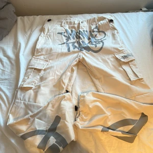 Beige cargopants med grått tryck - Säljer ett par beige cargopants med stora gråa graffiti-tryck framtill och på benen. Byxorna har flera fickor med lock och dragkedjor vid bensluten. Perfekta för dig som gillar streetwear och vill sticka ut.