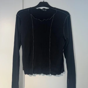 Svart ribbad långärmad tröja BikBok - Svart ribbad långärmad tröja från BikBok med kontrastsömmar i lila och vågig kant vid ärmslut och nederkant. Tröjan har en tight passform och är tillverkad i mjukt stretchigt material, perfekt för lager-på-lager eller som statement-piece. Passar s-L