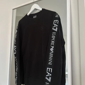 Svart EA7 Emporio Armani sweatshirt - Svart sweatshirt från EA7 Emporio Armani med vita logotexter längs båda ärmarna. Klassisk rund halsringning och långärmad modell. Perfekt för dig som gillar stilrena streetwear-plagg med tydlig branding.