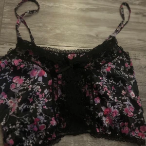 Blommig svart linnetopp med spets - Supersöt linnetopp i svart med blommigt mönster i rosa och lila toner. Toppen har tunna axelband, spetsdetaljer längs kanterna och en liten rosett framtill. Tillverkad i 100% polyester och har en lätt croppad passform. Perfekt för dig som gillar romantiska vibes.