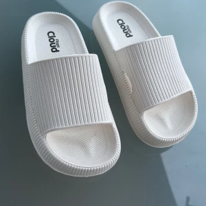 Vita Cloud Flops sandaler - Chunky vita sandaler från Cloud Flops med öppen tå och bred rem över foten. Tillverkade i mjuk syntet med ribbad struktur och mönstrad sula för extra grepp. Perfekta för chill dagar eller att glida runt i hemma eller på stranden. Det står storlek 36-37 under, men hon som använt dem har 38 och det funkat fint