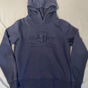 Mörkblå hoodie från GANT - Säljer en mörkblå hoodie från GANT med broderad logga och text på bröstet. Tröjan har huva, långa ärmar och ribbade muddar. Perfekt för dig som gillar klassisk och sportig stil. Materialet är mjukt och skönt i bomullsmix.