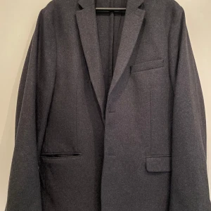 Mörkgrå kavaj från H&M, storlek 54 - Snygg mörkgrå ulliknande kavaj från H&M i klassisk modell med smala slag och tre fickor framtill. Kavajen har knappar vid ärmsluten och är tillverkad i ett mjukt material som ger en stilren look. Perfekt för dig som vill ha en enkel och tidlös stil.