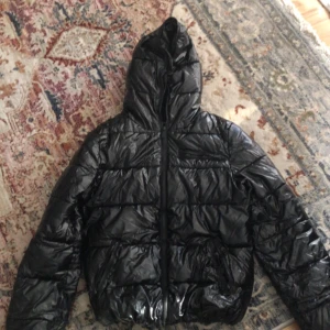 Svart glansig pufferjacka med huva - Säljer en svart pufferjacka med glansig finish och huva. Jackan har quiltad design, dragkedja framtill och långa ärmar. Perfekt för kalla dagar när du vill hålla stilen och värmen.