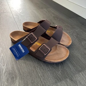 Bruna Birkenstock sandaler - Klassiska bruna Birkenstock sandaler med två breda remmar och justerbara metallspännen. Fotbädden är i mocka och sulan är i kork för extra komfort. Perfekta för varma dagar och enkel att matcha med allt. Endast testade en gång men passade inte mig