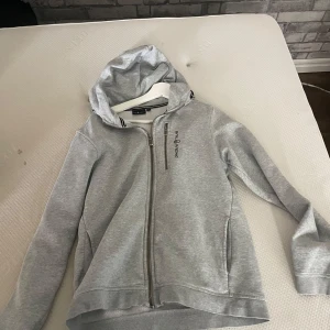 Grå hoodie från Sail Racing med dragkedja - Snygg ljusgrå hoodie från Sail Racing med hel dragkedja framtill och huva. Tröjan har en  logga på bröstet och praktiska fickor på sidorna. Perfekt för dig som gillar sportig och clean stil.