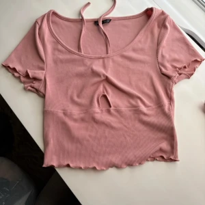 Rosa ribbad croppad topp med knyt - Supersöt ljusrosa croppad topp med ribbad struktur, korta ärmar och knytband i nacken. Toppen har en liten cut-out framtill och vågig kant nertill och vid ärmslut. 