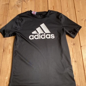 Svart Adidas t-shirt med logga - Svart t-shirt från Adidas med stor vit logga framtill. Tillverkad i lätt och sportig polyester, perfekt för träning eller chill. Klassisk passform med rund hals och korta ärmar. Snygg och enkel stil som funkar till det mesta.