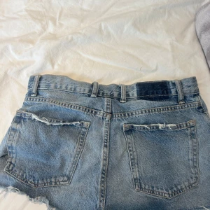 Blå slitna jeansshorts från Zara - Snygga blå jeansshorts från Zara med hög midja och råa, fransiga kanter. Shortsen har flera slitningar och knappar framtill för en cool vintagekänsla. Perfekta för sommardagar och festivaler. Klassisk femficksmodell med bälteshällor.