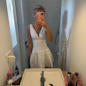 Vit kort sommarklänning med v-ringning - Supersöt vit kort klänning med djup v-ringning och smockad midja. Klänningen är ärmlös och har en luftig kjol som ger en riktigt fräsch sommarkänsla. Perfekt för varma dagar och enkel att styla med smycken eller sandaler.