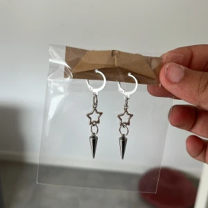 Silvriga örhängen med stjärna och spike - Snygga silvriga örhängen med stjärnformad detalj och hängande spike. Perfekta för dig som gillar en edgy och unik stil. Örhängena är i metall och har en cool design som sticker ut.