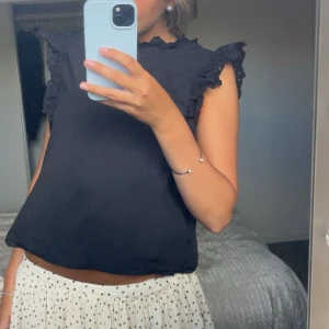 Svart blus med volangärmar - Supersöt svart blus med volangdetaljer på ärmarna och liten volang vid halsen. Ärmlös modell med broderade spetsdetaljer som ger en romantisk vibe. Perfekt att styla med kjol eller jeans för en trendig look.